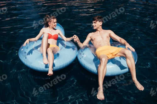 Preview: happy young couple holding hands while relaxing on swim rings in swimming pool
