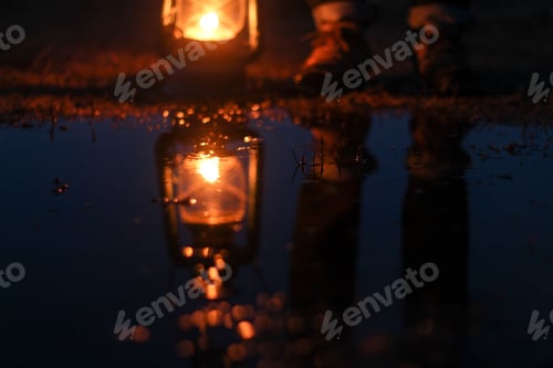 Preview: A person standing next to a lit lantern reflected on a puddle