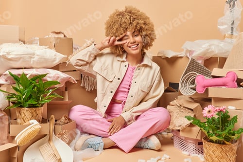 Preview: Happy young curly haired woman sits in lotus pose on floor makes peace gesture over eye enjoys start