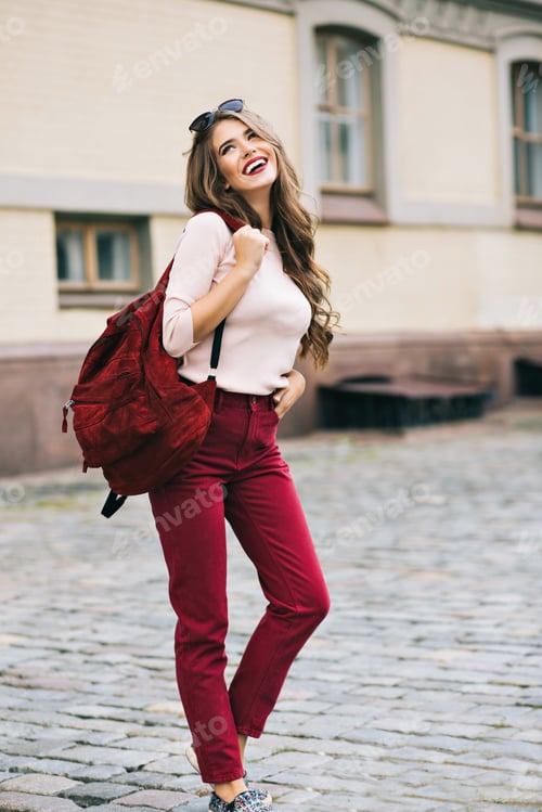 Preview: Fashion girl in vinous pants with bag and long hair is posing on pavement in city. She is smiling an