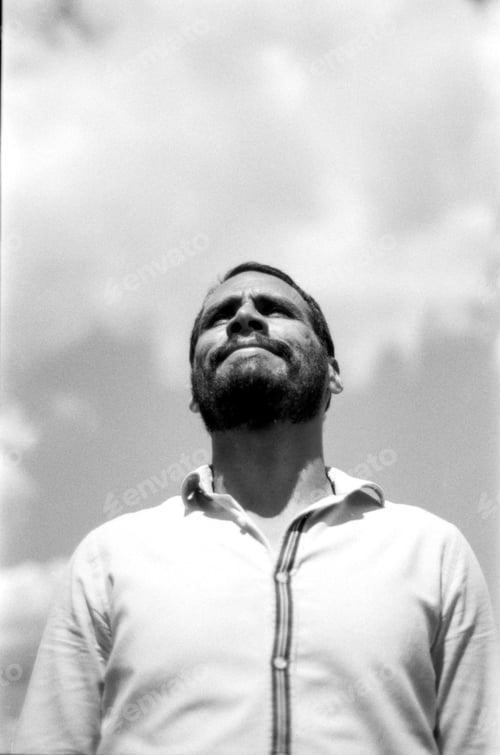 Preview: Bearded man looking up at cloudy sky in monochrome analog photo
