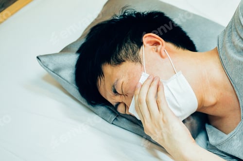 Preview: Young Asian man coughing and suffering in medical mask inside home bedroom - illness and fever