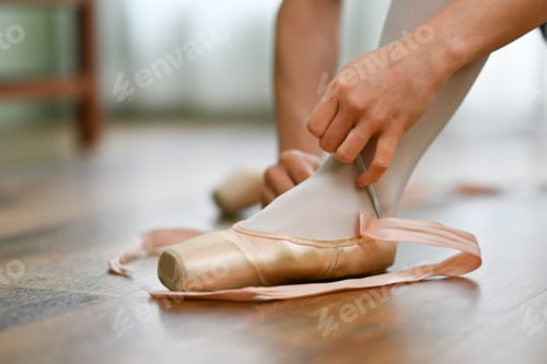 Preview: Young ballerina sitting on wooden floor and tying pointe shoes before training
