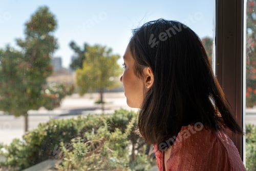 Preview: young woman looking out of the window, seeing a park and the sunny day