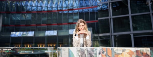 Preview: Beautiful young woman in beige suit, standing near office buildings in city center, smiling and
