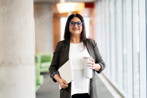 Preview: Mature Indian woman wearing eyeglasses holding digital tablet, bottle of water looking at camera