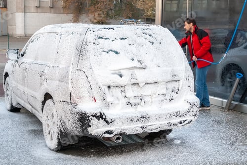Preview: A man in a red jacket covers a black car with foam at a self-service car wash. Back view