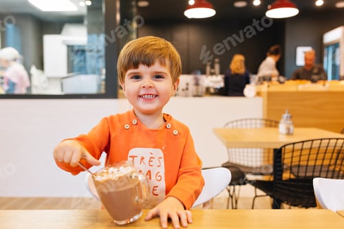Preview: Little child boy in orange sweater having fun with a cup of lot chocolate in cafe. He is smiling to