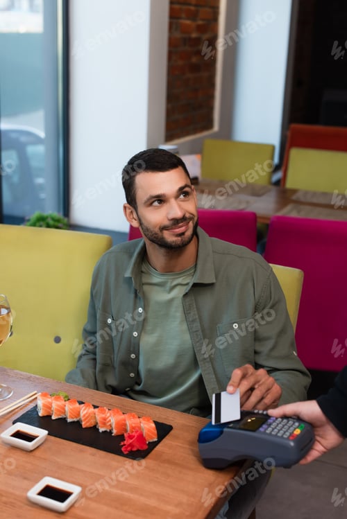 Preview: smiling man paying with credit card through payment terminal near tasty sushi