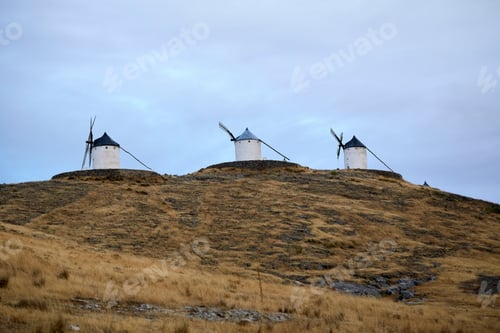 Preview: Three windmills on a grassy hilltop under a cloudy sky. Toledo, Spain