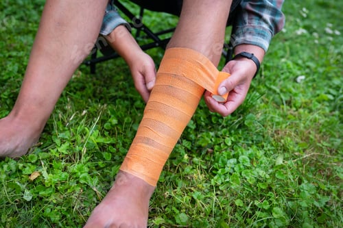 Preview: A person s leg being bandaged with an orange bandage. The person is sitting on a chair on a lawn