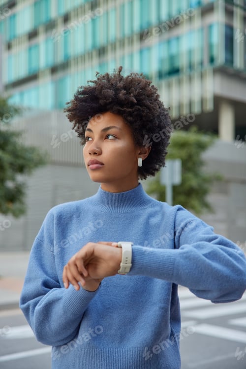 Preview: Outdoor shot of pensive curly haired Afro American woman waits for someone at street checks time onn