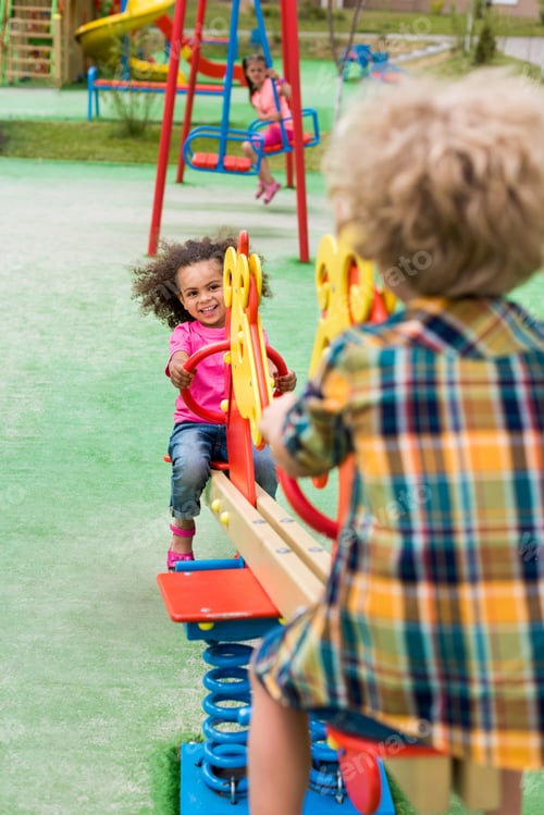 Preview: rear view of boy riding on rocking horse with smiling african american little kid at playground
