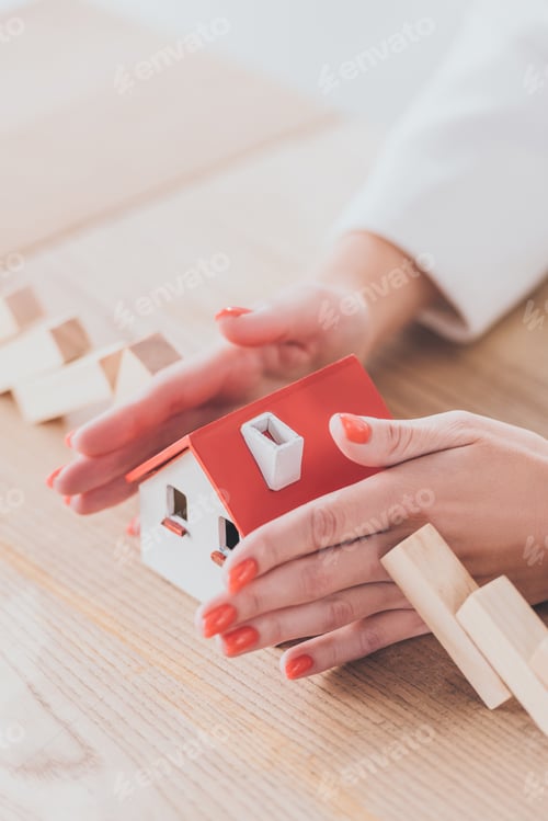 Preview: partial view of businesswoman protecting house model from falling wooden blocks with hands