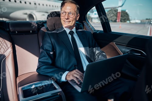 Preview: Cheerful businessman with laptop in car at airport