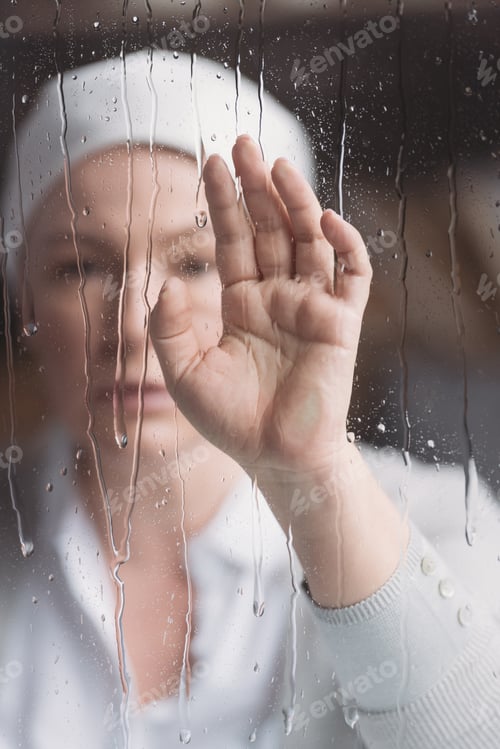 Preview: close-up view of sick mature woman in kerchief touching window with raindrops