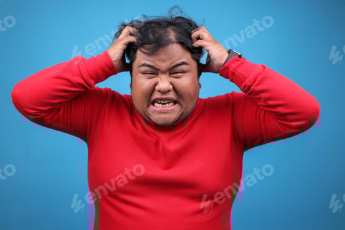 Preview: Young Fat Man Showing Stress, Hands Holding Head While Screaming Against Blue Studio Wall