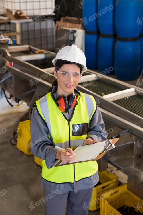 Preview: Female inspector inspecting conveyor in plant wearing helmet earmuffs glasses holding clipboard