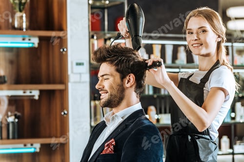Preview: beautiful young hairstylist smiling at camera while drying and combing hair to handsome client in