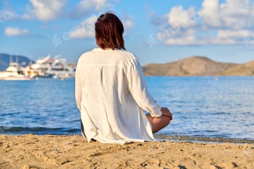Preview: Mature woman sitting in lotus position meditating on the beach, back view