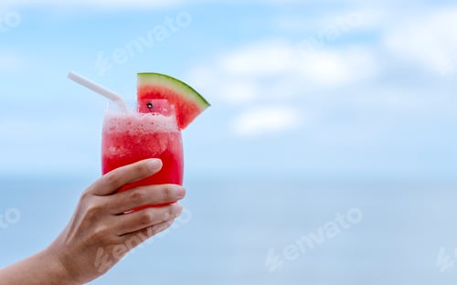 Preview: Closeup image of a hand holding a glass of watermelon smoothie by the sea with blue sky background
