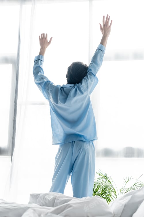 Preview: back view of man in blue pajamas stretching with raised hands near window