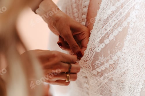 Preview: Young woman putting on a white lace wedding dress while being assisted by another woman