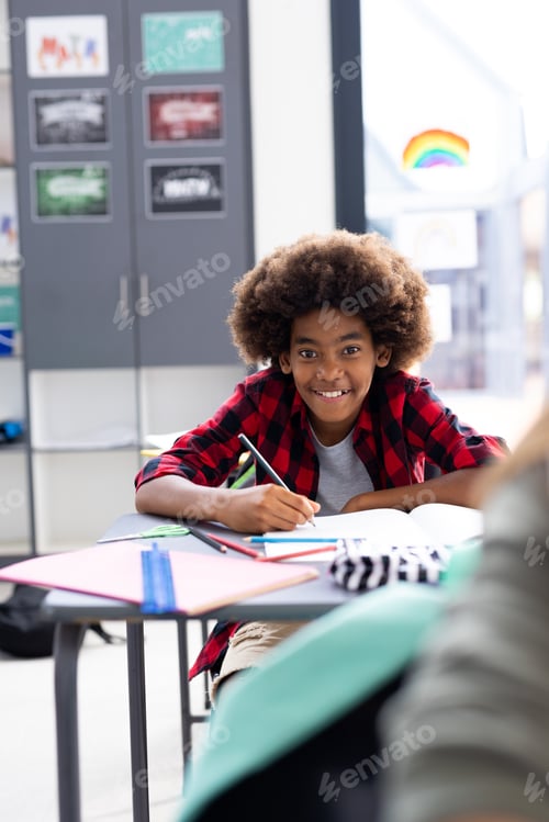 Preview: Vertical portrait of smiling african american boy with afro working at desk in classroom, copy space