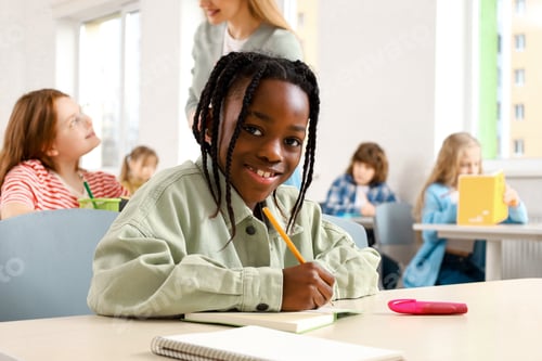 Preview: Happy African American boy studying sitting in classroom and writing in notebook, looking at camera