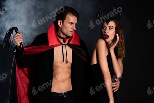Preview: handsome man in cloak holding flogging whip near surprised woman with horns on black with smoke