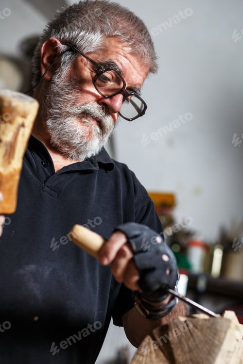 Preview: Senior sculptor working on his wood sculpture in his workshop with hammer and chisel.