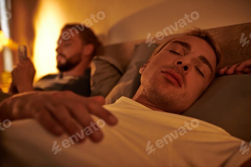 Preview: gay man sleeping near unfaithful boyfriend browsing date app on smartphone at night in bedroom