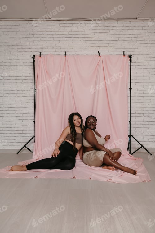 Preview: Two happy plus size African women in sportswear sitting and bonding on pink fabric