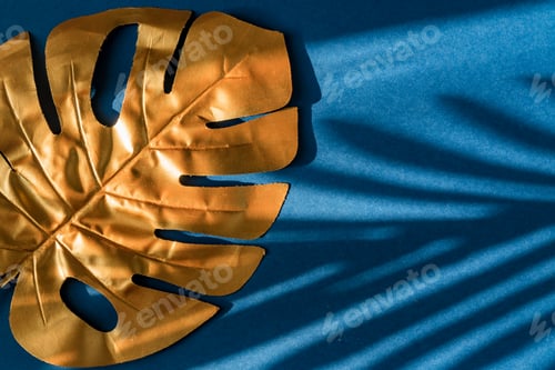 Preview: Stylish gold coloured monstera leaf on blue background with shadows