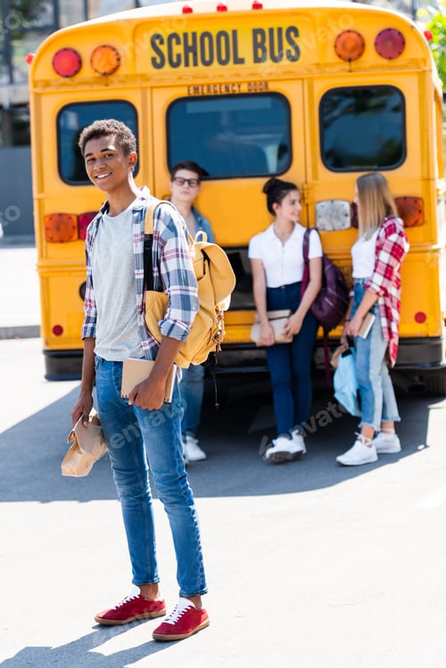 Preview: handsome smiling teen schoolboy looking at camera while his classmates standing behind near school