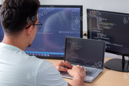Preview: A young programmer wearing glasses working with three monitors, writing programming code.