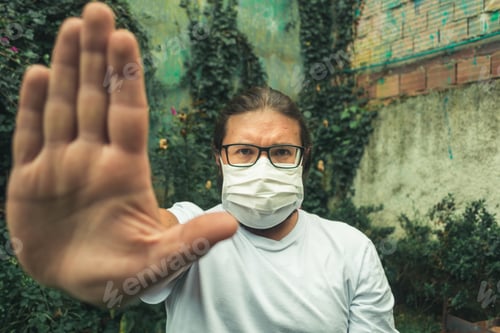 Preview: Man in glasses wearing a white face mask to protect from dust and coronavirus showing stop sign