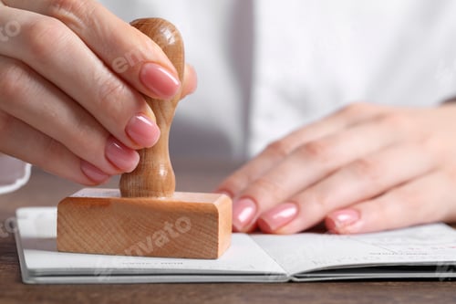 Preview: Ukraine, Lviv - September 6, 2022: Woman stamping visa page in passport at wooden table, closeup