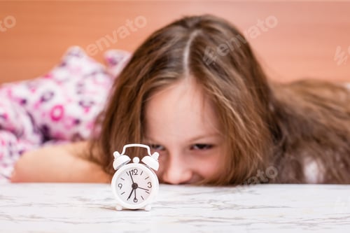 Preview: White alarm clock stands on the table on the background of a waking up girl