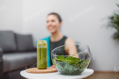 Preview: A young woman drinks a vegetable and fruit smoothie in her living room and doing yoga on yoga mat.