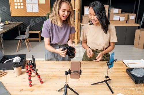 Preview: Girl holding photocamera over table while explaining colleague how to use it