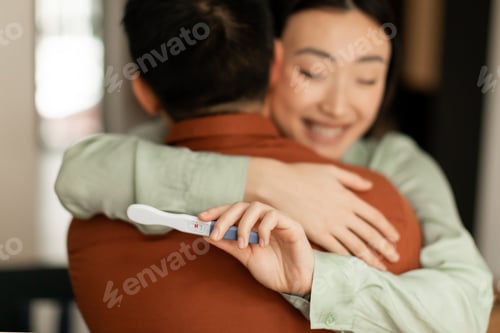 Preview: Happy young asian woman holding positive pregnancy test and hugging husband, loving family embracing