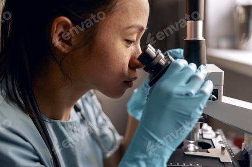 Preview: Asian woman in blue gloves looks into microscope lens sitting at workplace in laboratory