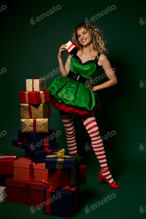 Preview: smiley elf in green dress with striped stockings posing with presents on dark green backdrop