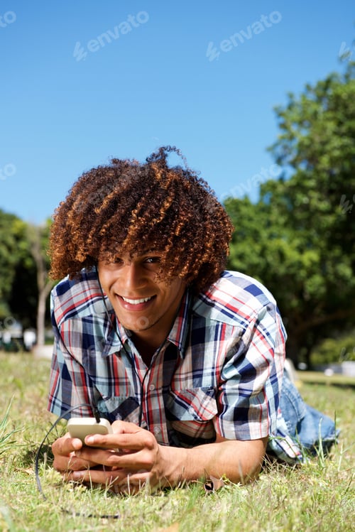 Preview: smiling young afro man lying down on grass in park holding phone