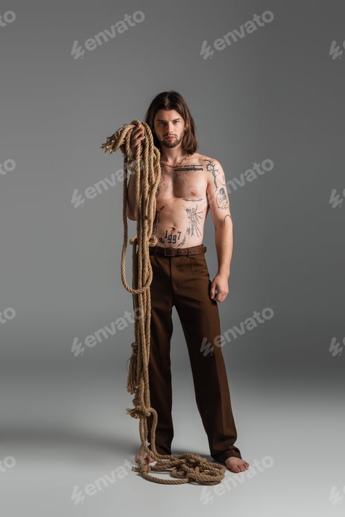 Preview: Barefoot and tattooed model holding rope on grey background