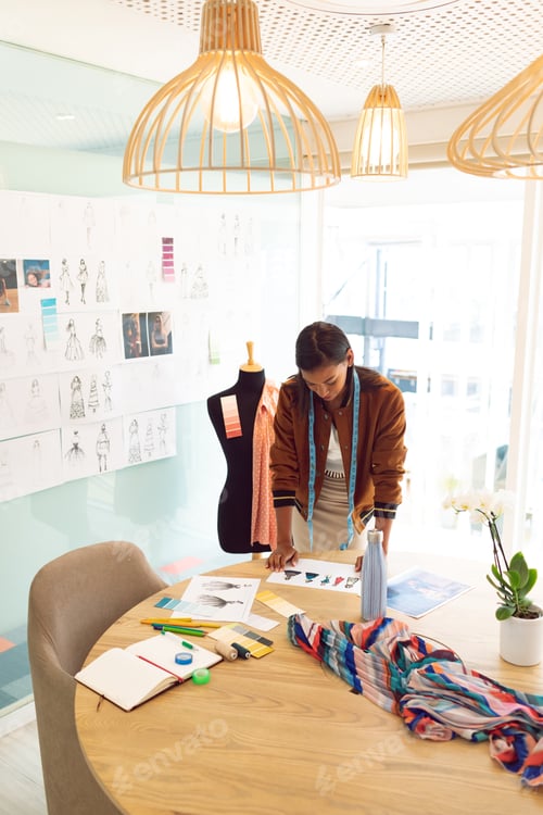 Preview: Mixed race female fashion designer looking at sketches on table
