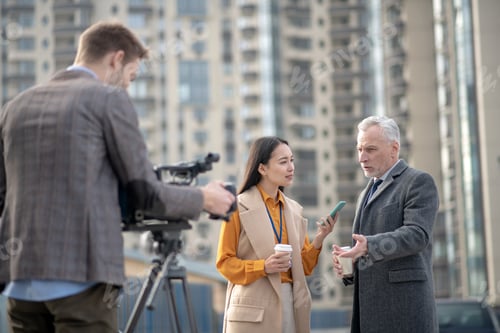Preview: Female reporter in beige outfit asking questions to a grey-haired man