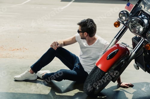 Preview: Stylish young man in sunglasses smoking while sitting near motorcycle and looking away