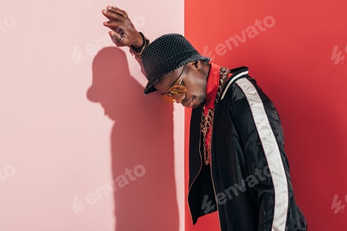 Preview: stylish african american man in hat, sunglasses and jewellery looking down in studio
